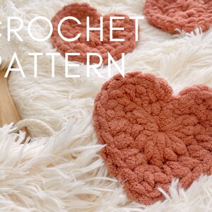 May include: Three crocheted heart-shaped coasters in a dusty rose color on a white fluffy surface. The text "CROCHET PATTERN" is written in white on the surface.
