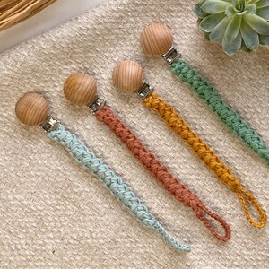 May include: Four crocheted pacifier clips with wooden beads and metal clips. The clips are in shades of blue, green, orange, and brown.