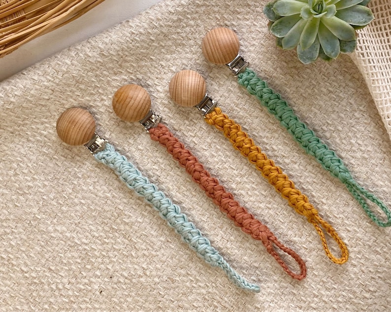 May include: Four crocheted pacifier clips with wooden beads and metal clips. The clips are in shades of blue, green, orange, and brown.
