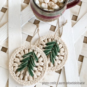 Puede incluir: Dos adornos navide&ntilde;os de ganchillo en forma de &aacute;rbol de Navidad. Los adornos son de color beige con detalles de hilo verde. Los adornos est&aacute;n sobre una superficie blanca con un patr&oacute;n tejido.