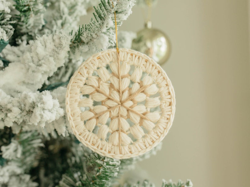 Puede incluir: Un adorno de copo de nieve de crochet blanco colgado de un &aacute;rbol de Navidad nevado.