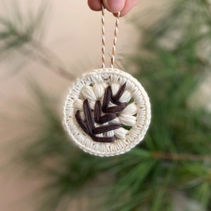 Puede incluir: Un adorno de crochet blanco con un dise&ntilde;o de hoja marr&oacute;n. El adorno est&aacute; colgado de una cuerda marr&oacute;n y blanca.