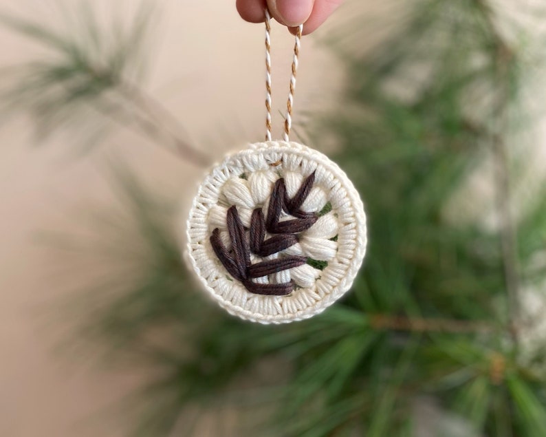 Puede incluir: Un adorno de crochet blanco con un dise&ntilde;o de hoja marr&oacute;n. El adorno est&aacute; colgado de una cuerda marr&oacute;n y blanca.