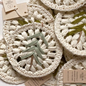 Puede incluir: Primer plano de varios adornos de ganchillo hechos a mano con un dise&ntilde;o de bordado verde y marr&oacute;n. Los adornos est&aacute;n hechos con fibras naturales y tienen una est&eacute;tica r&uacute;stica y de granja. El texto de la etiqueta dice "Christina Ann Studios Thoughtfully Handmade Goods for the Home Made in Kentucky".