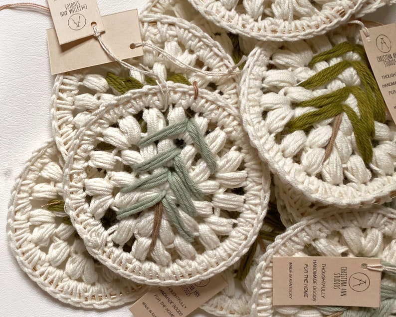 Puede incluir: Primer plano de varios adornos de ganchillo hechos a mano con un dise&ntilde;o de bordado verde y marr&oacute;n. Los adornos est&aacute;n hechos con fibras naturales y tienen una est&eacute;tica r&uacute;stica y de granja. El texto de la etiqueta dice "Christina Ann Studios Thoughtfully Handmade Goods for the Home Made in Kentucky".