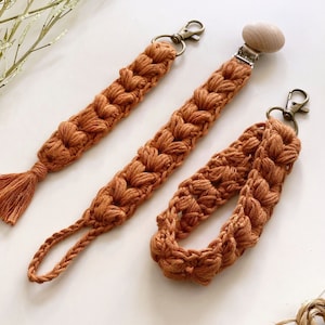 May include: Three crocheted cords in a burnt orange color. One cord has a wooden pacifier clip attached, another has a tassel, and the third has a key ring.
