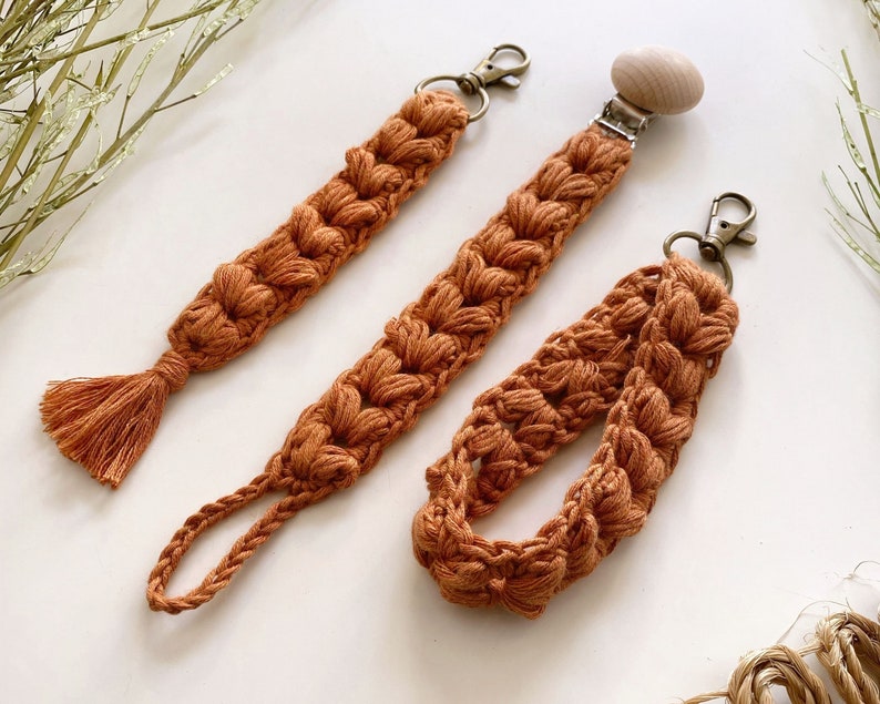 May include: Three crocheted cords in a burnt orange color. One cord has a wooden pacifier clip attached, another has a tassel, and the third has a key ring.