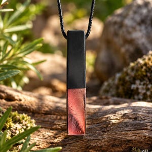 K&ouml;nnte beinhalten: Ein rechteckiger Anh&auml;nger mit zweifarbigem Design, bestehend aus einem mattschwarzen oberen Teil und einem metallisch kupferfarbenen unteren Teil. Der Anh&auml;nger h&auml;ngt an einer d&uuml;nnen schwarzen Kordel und hat eine klare, geometrische Form.