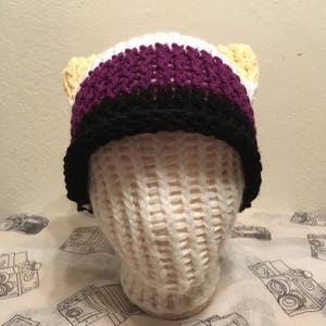 May include: A crocheted beanie hat with yellow, white, purple, and black stripes. The hat has two yellow cat ears.
