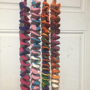 May include: Four colorful crochet spiral ornaments hanging from silver clips. Each spiral features a unique color combination, including pink, blue, orange, and purple. The ornaments are suspended in front of a white door, with tassels at the bottom.