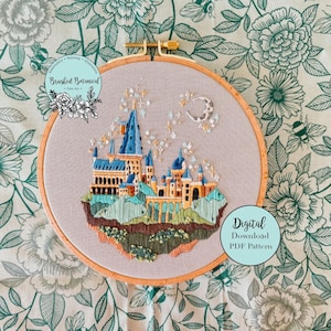 May include: Embroidered hoop art featuring a detailed depiction of Hogwarts castle with a blue and gold colour scheme. The castle is surrounded by a green landscape and a crescent moon with stars. The text "Digital Download PDF Pattern" is visible in the lower right corner.
