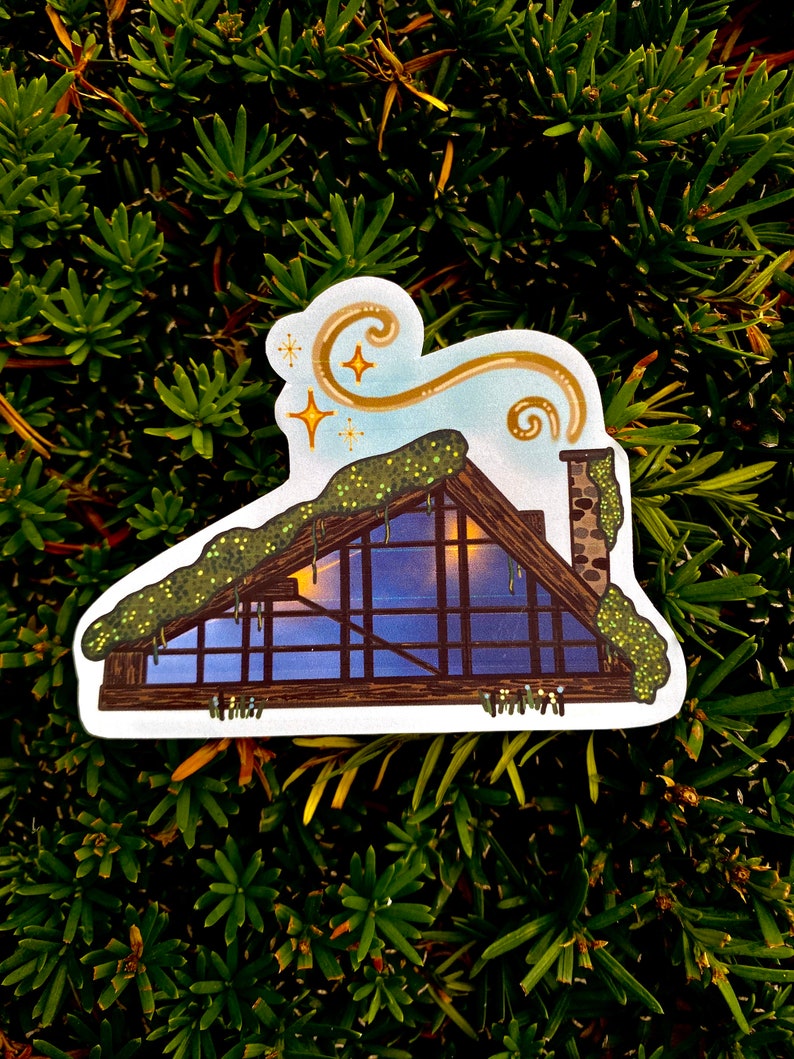 May include: A sticker featuring a whimsical illustration of a cabin with a chimney, a moss-covered roof, and a starry sky. The cabin has large windows and a warm light emanating from within. The sticker is set against a backdrop of green foliage.