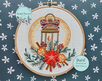 May include: Embroidered hoop art featuring a lantern with a red and white bow, surrounded by greenery, berries, and cinnamon sticks. The hoop is on a blue background with white flowers. The text "Digital Download PDF Pattern" is in a white circle on the bottom right.