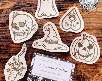 May include: A collection of Halloween-themed embroidery designs, including a skull, ghost, witch's hat, Ouija board, pumpkin, and broom. The designs are printed on fabric with a neutral color palette. A package labeled "Stick and Stitch" is also visible.