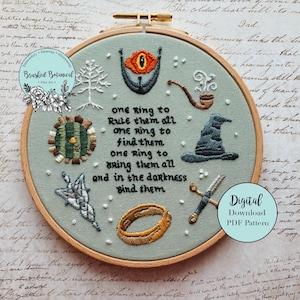 May include: Embroidered hoop art with a fantasy theme. The design includes an eye, a tree, a pipe, a hat, a sword, a ring, and text that reads "one ring to rule them all." The artwork is on a light green background, set in a wooden hoop.