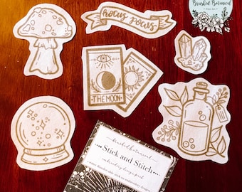 May include: A set of iron-on embroidery designs featuring a mushroom, a crystal, a bottle, a tarot card, a magic eight ball, and a banner that says "Hocus Pocus".