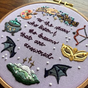 May include: Embroidered hoop art with a purple background and various colourful symbols including a cauldron, a paint palette, a feather, a mask, a bow and arrow, and bats. The text reads "To the stars who listen to the dreams that are answered."