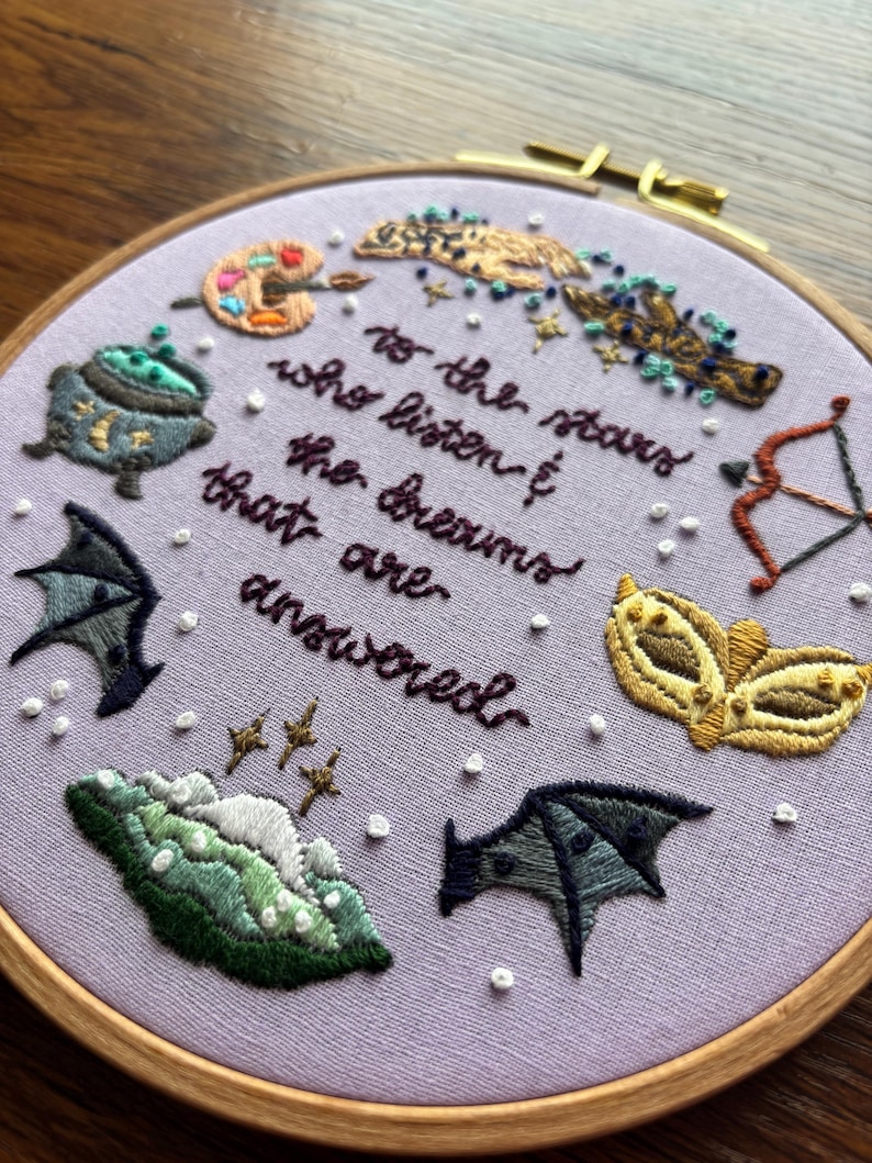May include: Embroidered hoop art with a purple background and various colourful symbols including a cauldron, a paint palette, a feather, a mask, a bow and arrow, and bats. The text reads "To the stars who listen to the dreams that are answered."