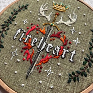 K&ouml;nnte beinhalten: Gesticktes Bild im Holzrahmen mit dem Wort "fireheart", Schwert, Krone und Geweih. Das Design zeigt rote und orangefarbene Flammen, wei&szlig;e Sterne und gr&uuml;nes Laub auf einem olivgr&uuml;nen Stoff. Der Rahmen hat einen Durchmesser von ca. 20 cm.