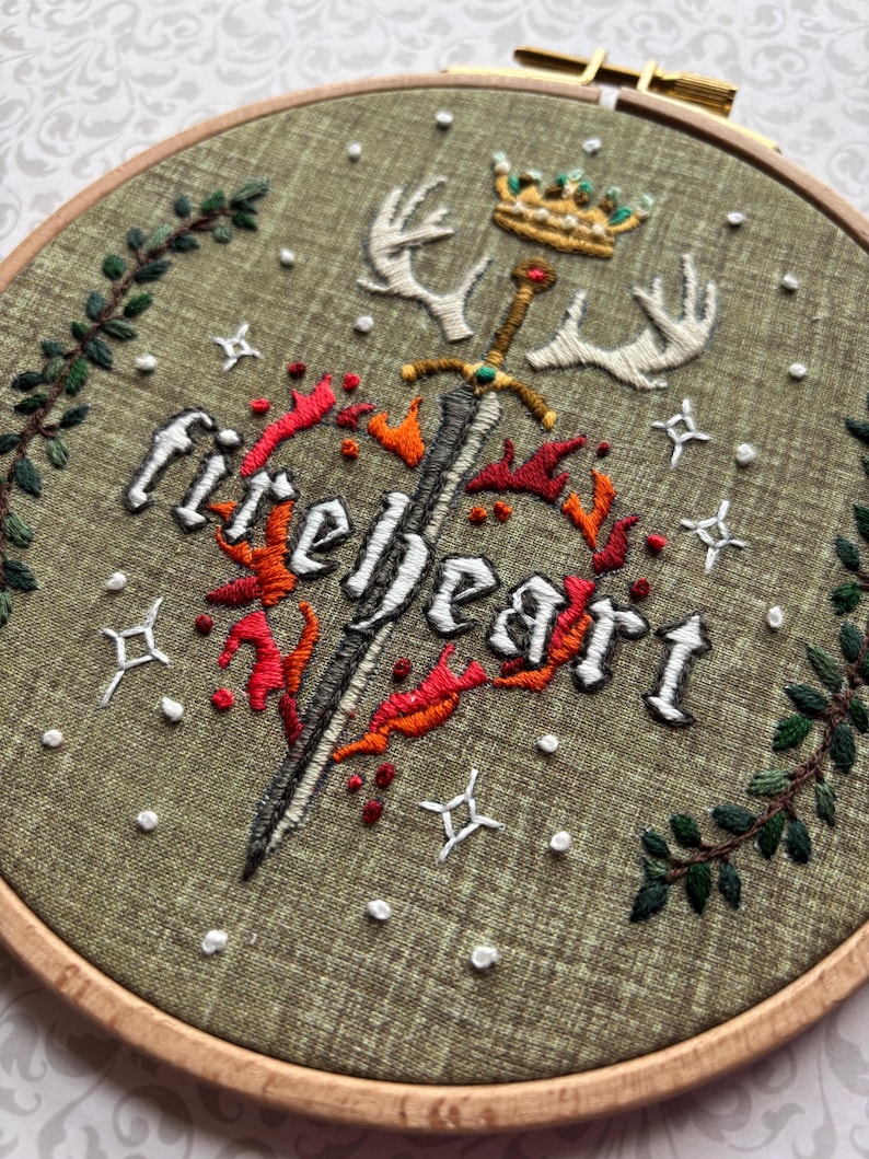 K&ouml;nnte beinhalten: Gesticktes Bild im Holzrahmen mit dem Wort "fireheart", Schwert, Krone und Geweih. Das Design zeigt rote und orangefarbene Flammen, wei&szlig;e Sterne und gr&uuml;nes Laub auf einem olivgr&uuml;nen Stoff. Der Rahmen hat einen Durchmesser von ca. 20 cm.
