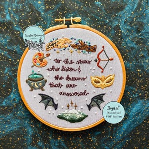 May include: Embroidered hoop art with a white background and colourful embroidery. The embroidery includes a cauldron, a paint palette, a bow and arrow, a mask, and wings. The text reads "to the stars who listen & the dreams that are answered".
