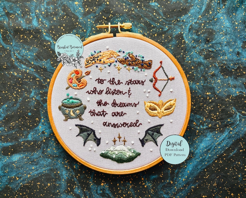 May include: Embroidered hoop art with a white background and colourful embroidery. The embroidery includes a cauldron, a paint palette, a bow and arrow, a mask, and wings. The text reads "to the stars who listen & the dreams that are answered".