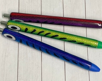 May include: Three pens with a fish design. The pens are red, green, and blue with glitter and black stripes. The pens have a black eye and a white pupil.
