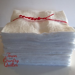 May include: A stack of white cotton batting squares tied together with a red ribbon. The batting is labeled "Town and Country Quilter".