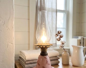 Peut inclure: Une lampe de table de style vintage avec une base en verre rose pâle ornée de motifs de vigne en relief, un col en laiton et une cheminée en verre transparent. La lampe émet une lueur chaude depuis une source lumineuse interne.