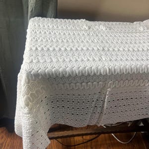 May include: A white, textured crochet blanket draped over a wooden table. The blanket features a repeating wave-like pattern, creating a visually interesting surface. The blanket's edges hang over the table, showcasing the intricate design.