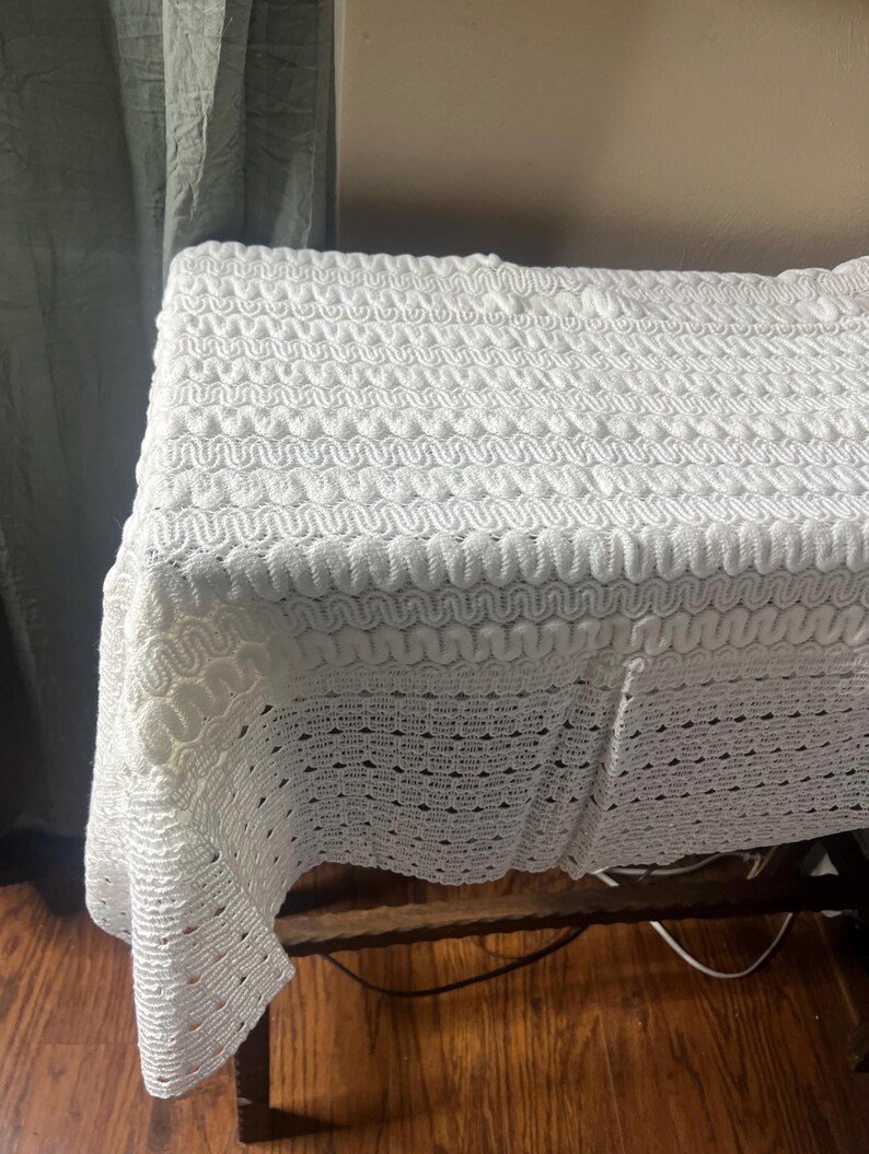 May include: A white, textured crochet blanket draped over a wooden table. The blanket features a repeating wave-like pattern, creating a visually interesting surface. The blanket's edges hang over the table, showcasing the intricate design.