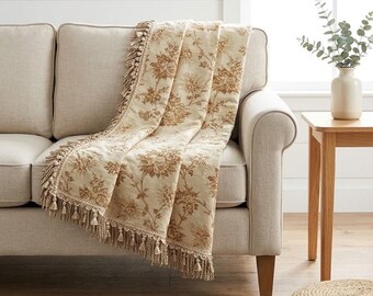 Peut inclure: Un jeté de canapé couleur crème orné d'un motif floral marron et d'une bordure à franges décoratives. Le tissu présente un dessin botanique détaillé avec des feuilles et des fleurs complexes, drapé sur un canapé aux tons neutres.