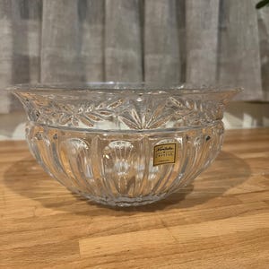 May include: A clear crystal bowl with a decorative leaf pattern around the rim and vertical ridges on the sides. The bowl has a wide, shallow shape and a small rectangular label that reads "Noritake Crystal GERMANY".