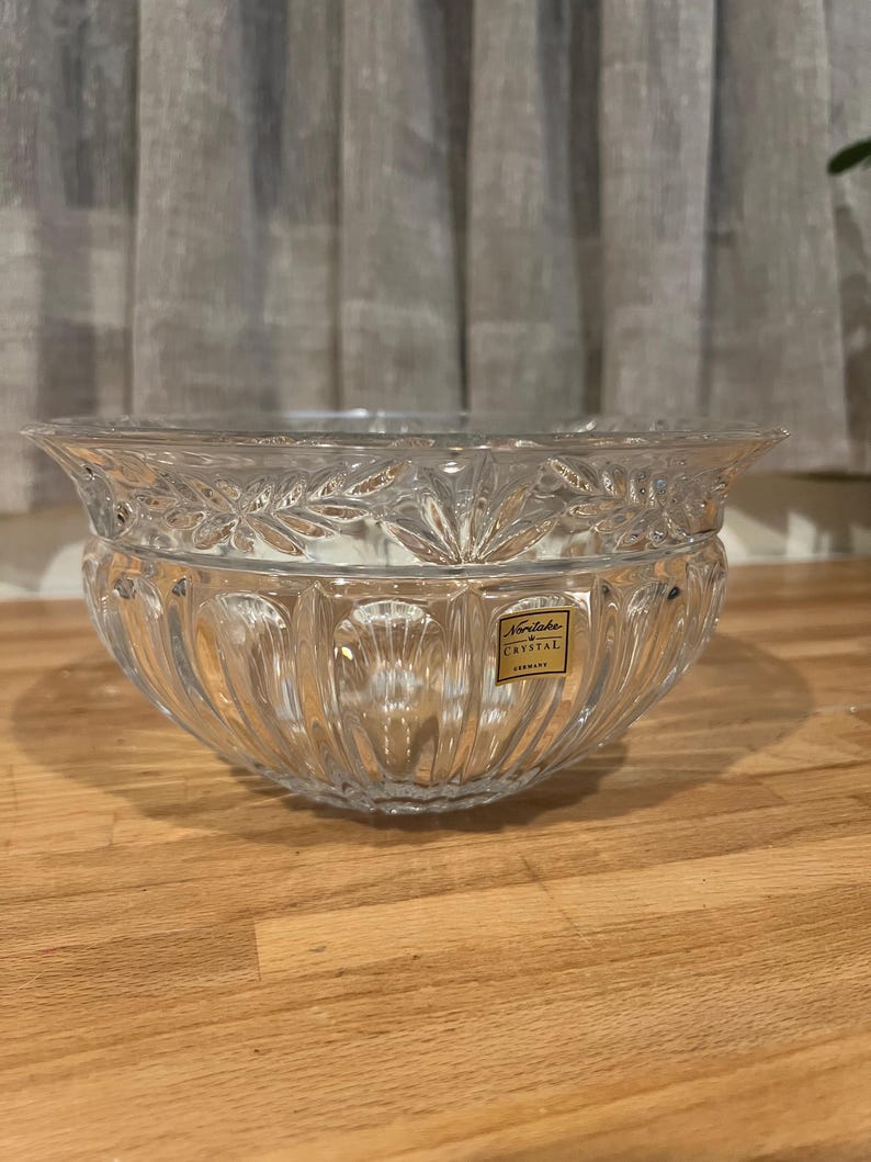 May include: A clear crystal bowl with a decorative leaf pattern around the rim and vertical ridges on the sides. The bowl has a wide, shallow shape and a small rectangular label that reads "Noritake Crystal GERMANY".