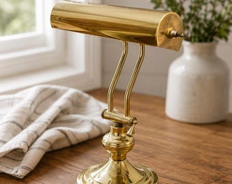 Peut inclure: Une lampe de piano en laiton poli dotée d'un abat-jour cylindrique, d'un col réglable à deux bras et d'une base circulaire. La lampe comprend un petit bouton sur le côté de l'abat-jour et un fin cordon d'alimentation sortant de la base.