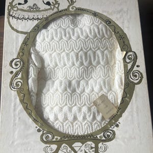 May include: A vintage white knitted garment, possibly a sweater or top, displayed within a decorative oval frame on a box. The box has an ornate design with gold and black accents. A small tag is attached to the garment.