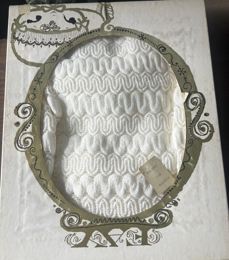May include: A vintage white knitted garment, possibly a sweater or top, displayed within a decorative oval frame on a box. The box has an ornate design with gold and black accents. A small tag is attached to the garment.