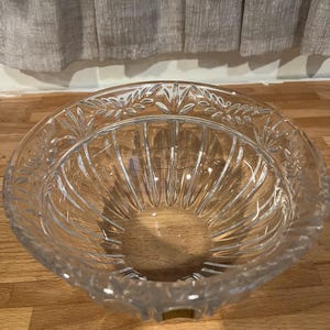 May include: A clear, decorative glass bowl with a wide, shallow design. The bowl features a detailed leaf pattern around the rim and vertical grooves on the interior. The bowl is sitting on a wooden surface.