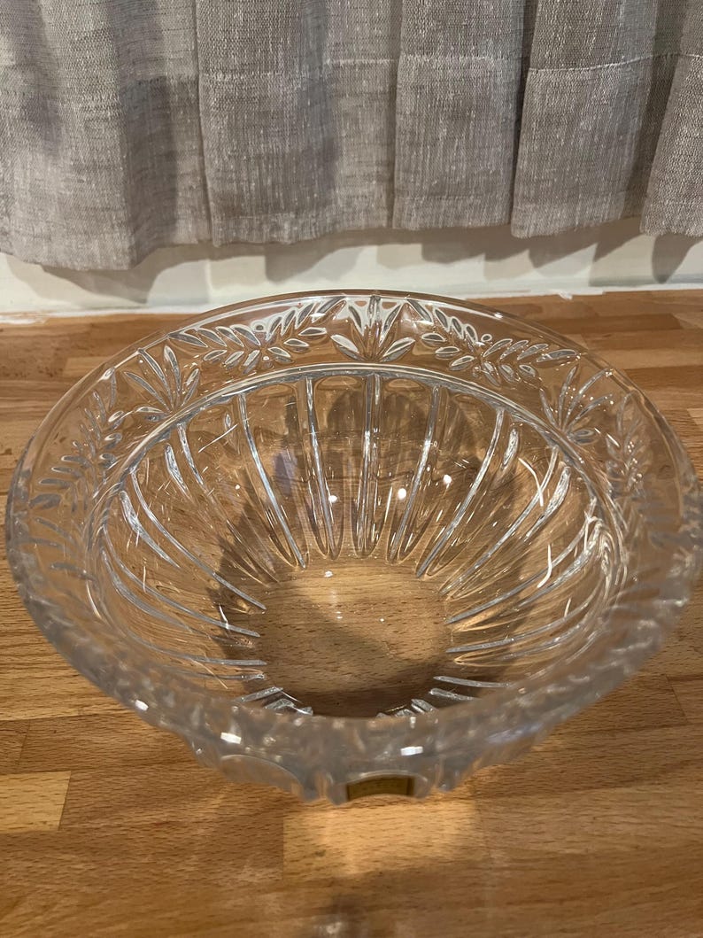 May include: A clear, decorative glass bowl with a wide, shallow design. The bowl features a detailed leaf pattern around the rim and vertical grooves on the interior. The bowl is sitting on a wooden surface.