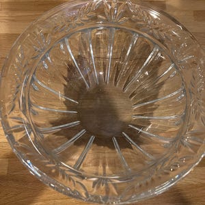 May include: A clear glass bowl with a decorative leaf pattern around the rim. The bowl has a circular shape with vertical lines radiating from the center. The bowl is sitting on a wooden surface.