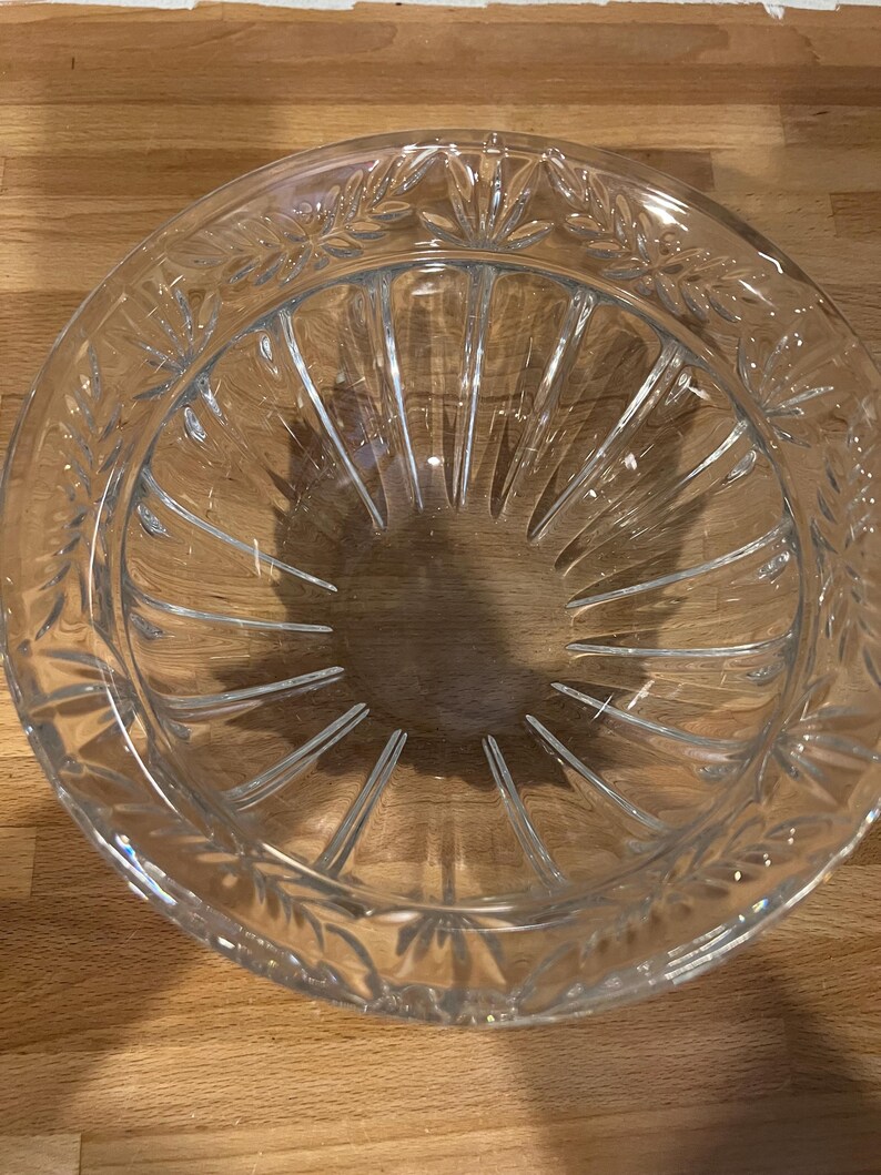 May include: A clear glass bowl with a decorative leaf pattern around the rim. The bowl has a circular shape with vertical lines radiating from the center. The bowl is sitting on a wooden surface.