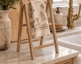 Peut inclure: Un porte-serviettes en bois clair avec un design en forme de A, supportant un torchon de cuisine crème orné d'un motif de feuilles botaniques vertes et d'un bord effiloché. Le support possède deux barres horizontales et quatre pieds.