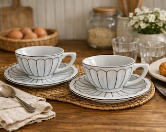 Peut inclure: Deux tasses à thé et soucoupes en céramique blanche avec une bordure festonnée noire sont présentées sur un tapis tissé. Une cuillère et une serviette en lin sont au premier plan. La scène suggère un petit-déjeuner.