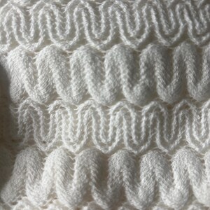 May include: Close-up of a white knitted fabric with a textured, wavy pattern. The design features repeating, rounded zigzags, creating a three-dimensional effect. The soft, off-white color suggests a cozy, handmade textile, possibly for a blanket or scarf.