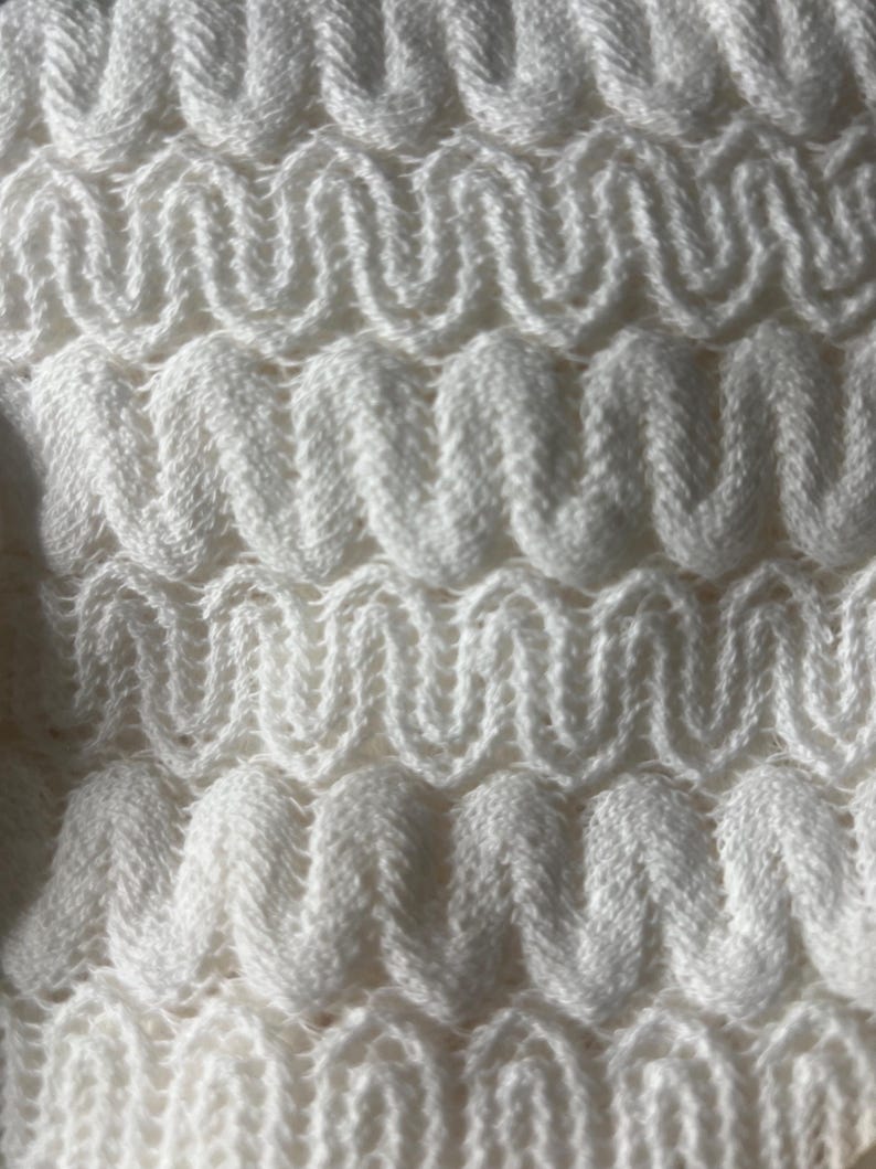 May include: Close-up of a white knitted fabric with a textured, wavy pattern. The design features repeating, rounded zigzags, creating a three-dimensional effect. The soft, off-white color suggests a cozy, handmade textile, possibly for a blanket or scarf.