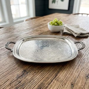 May include: A rectangular silver-toned serving tray with rounded corners and ornate, curved handles on each side. The surface of the tray features an intricate, embossed floral and scrollwork pattern with a smooth, plain center area.
