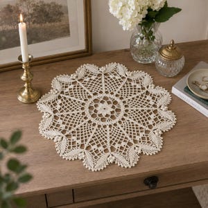 Könnte beinhalten: Eine runde, cremeweiße Häkeldeckchen mit einem komplizierten, durchbrochenen geometrischen Muster, einem zentralen Blumenmotiv und einem gewellten Rand, der mit kleinen, erhabenen Maschengruppen verziert ist, was ein zartes, kreisförmiges Textildesign ergibt.