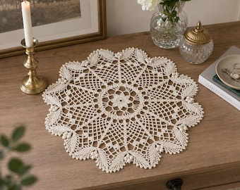May include: A round, off-white crochet doily featuring an intricate, openwork geometric pattern with a central floral motif and scalloped edges decorated with small, raised clusters of stitches, creating a delicate, circular textile design.