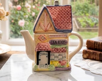 May include: A house-shaped ceramic teapot with a red brick roof and a sign saying "TEA ROOM." The teapot has a cream-coloured spout and handle, with colourful details on the walls and trim. It is placed on a marble surface.