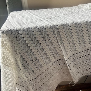 May include: A white, intricately patterned lace tablecloth. The tablecloth is draped over a table, showcasing a detailed, openwork design. The lace features a repeating, wave-like pattern, adding texture and visual interest to the textile.